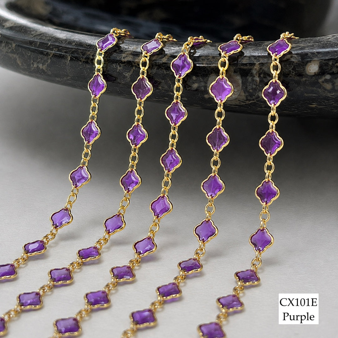 Five strands of purple gemstone chain jewelry with gold links, draped over a dark stone surface on a light gray background. A label in the bottom right reads &
