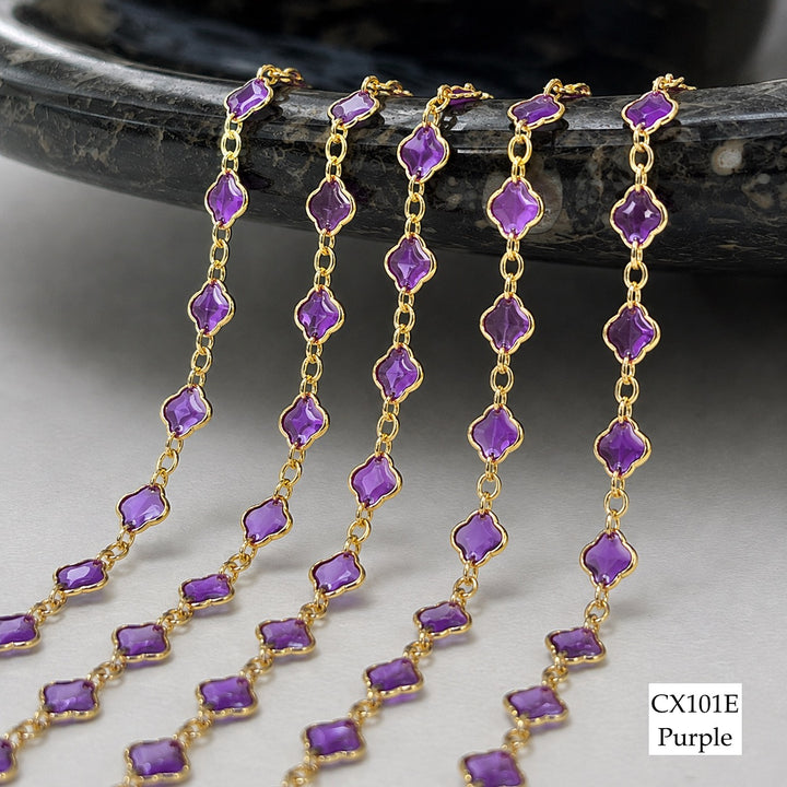 Five strands of purple gemstone chain jewelry with gold links, draped over a dark stone surface on a light gray background. A label in the bottom right reads &