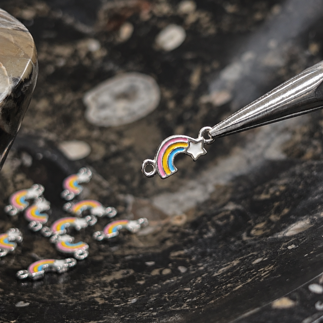 A small silver rainbow and star charm is held by metal tweezers over a dark marbled surface, with several matching charms scattered nearby.