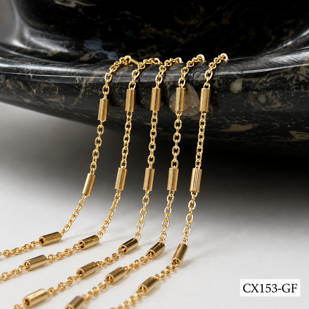 A close-up of a gold chain necklace with cylindrical barrel links, draped over a dark polished stone surface on a white background.