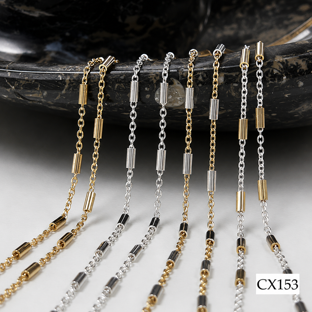 Six delicate chain necklaces in gold and silver tones with cylindrical and bead links, arranged on a dark polished stone slab over a light surface. A small label reading CX153 appears in the bottom right corner.