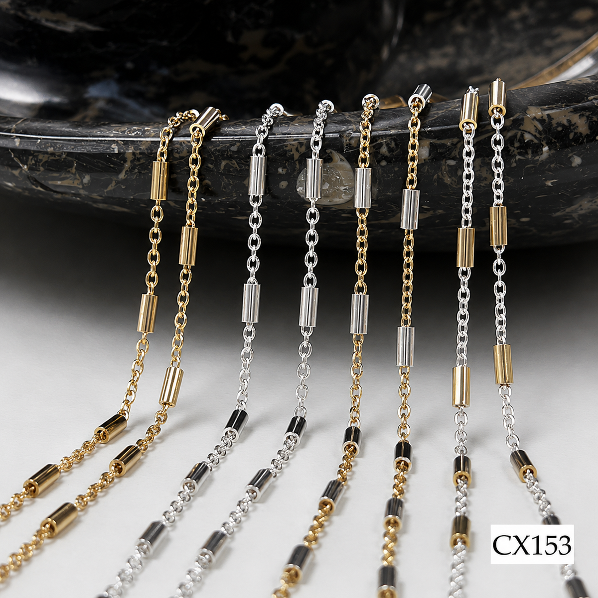 Six delicate chain necklaces in gold and silver tones with cylindrical and bead links, arranged on a dark polished stone slab over a light surface. A small label reading CX153 appears in the bottom right corner.